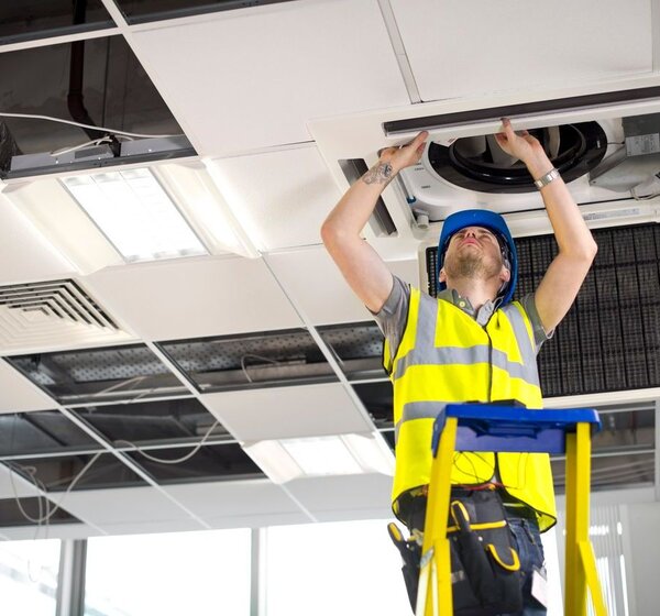 Image showing an electrician on a ladder fitting air conditioning unit into a ceiling. In a bright office an engineer in a hard hat stands on a ladder looking up as he fits an air conditioning unit into the ceiling.