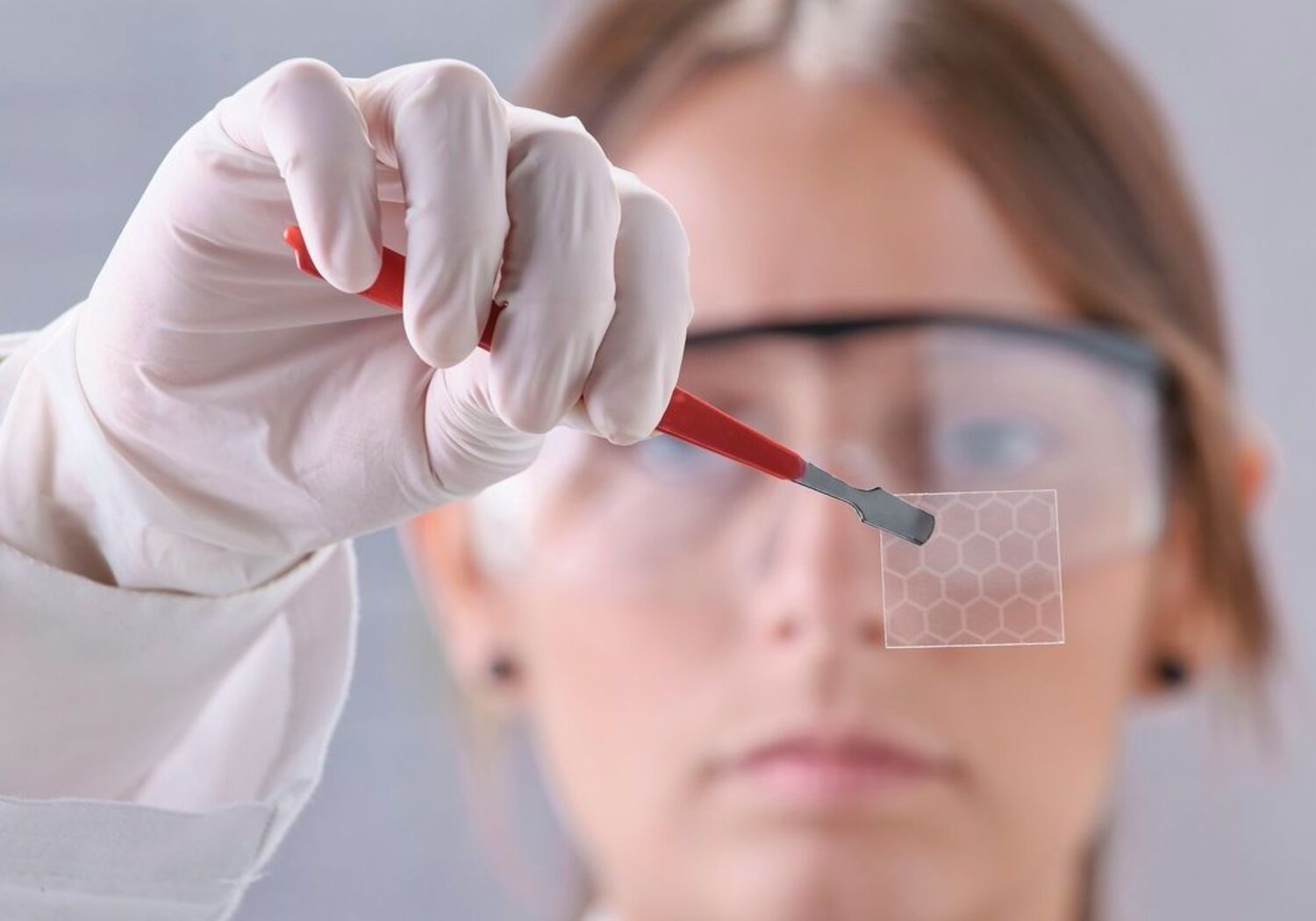 Image showing a scientist holding a hexagonal graphene molecule.Close up of a scientist’s hand holding a pair of forceps between which is held a transparent square of hexagonal graphene.