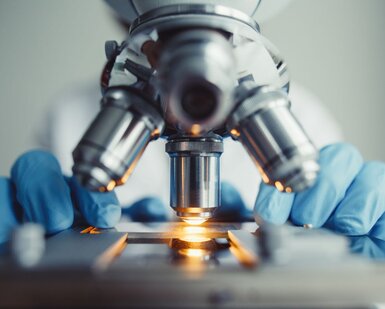 In the centre of the picture is a microscope with 4 lenses. Behind and out of site, sits a scientist with only gloved hands showing either side of the microscope