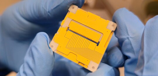 Image showing a pair of blue gloved hands holding the new 3D ion trap, which resembles a yellow square 3 cm by 3 cm