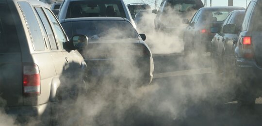 A photograph of a number of cars stopped at a traffic slight. The image is hazy with exhaust fumes coming from the vehicles’ exhausts