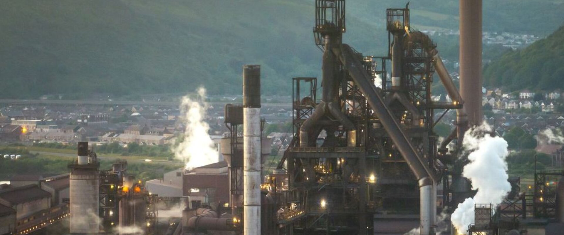 Against a hazy grey sky a large steel plant sits in a valley with white smoke plumes emitted from its large chimney stacks