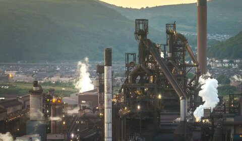 Against a hazy grey sky a large steel plant sits in a valley with white smoke plumes emitted from its large chimney stacks