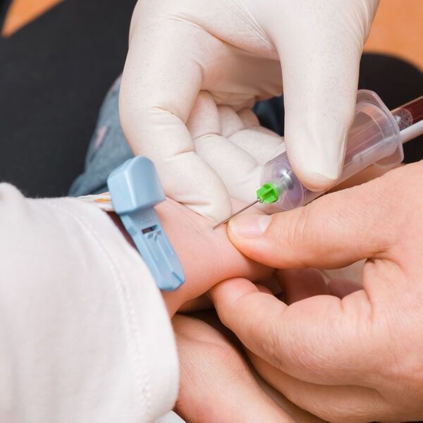 A close-up image of the hands of a doctor with white rubber gloves taking blood out of a young child’s arm with a syringe