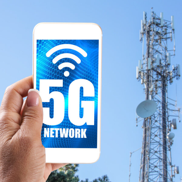 In the background is a high telecommunication tower with dishes and in the foreground a hand holds a mobile phone with the words “5G network” on the screen