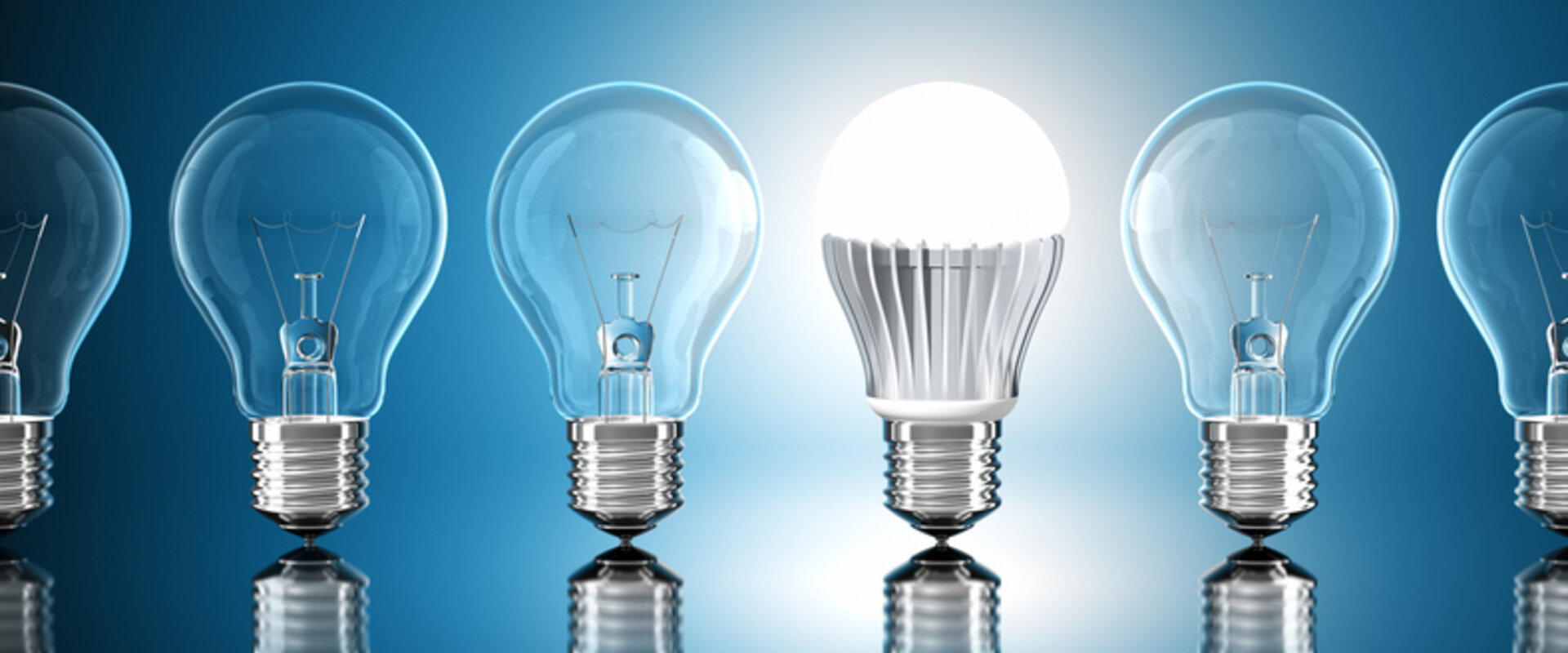 Against a pale blue background is a row of unlit incandescent screw light bulbs with one lit LED bulb