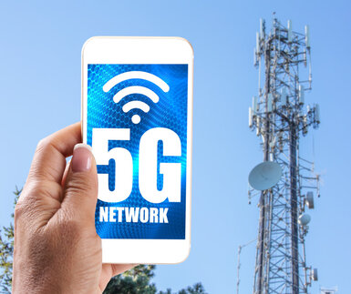 In the background is a high telecommunication tower with dishes and in the foreground a hand holds a mobile phone with the words “5G network” on the screen