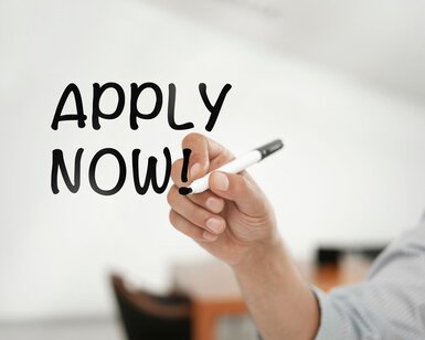 A hand writing “APPLY NOW!” on a whiteboard