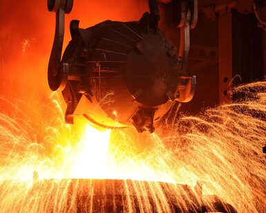 In a factory environment an overhead metal bucket pours orange glowing, molten steel into a vessel below