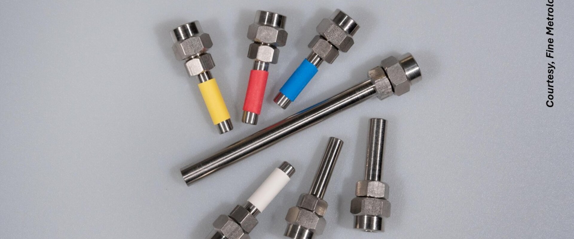 Against a grey background are seven different types of WaferCal permeation tubes from the company Fine Metrology. These resemble “nuts and bolts” with different coloured bands on the “bolt” of the tubes