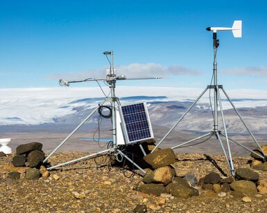 On a rocky ground in the mountains and framed by a blue sky sits a meteorological station. It is composed of a small monitor with a solar panel on the left and on the right a weathervane