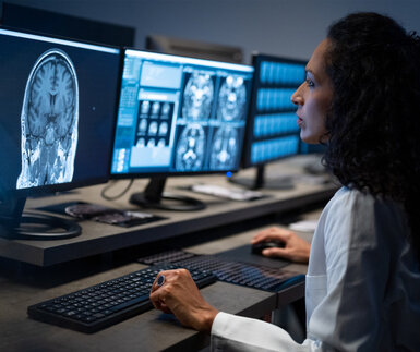 A radiologist sits in a darkened room inspecting MRI images of a patient’s head on a computer screen