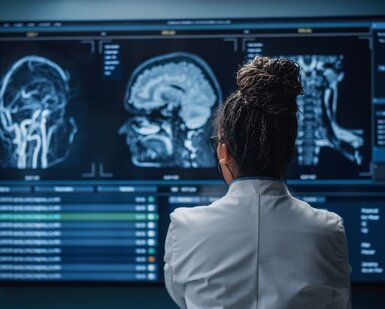 In the foreground is the back of a female neuroscientist in a white lab coat and in the background a wall of MRI scans of a patient’s brain that she is studying