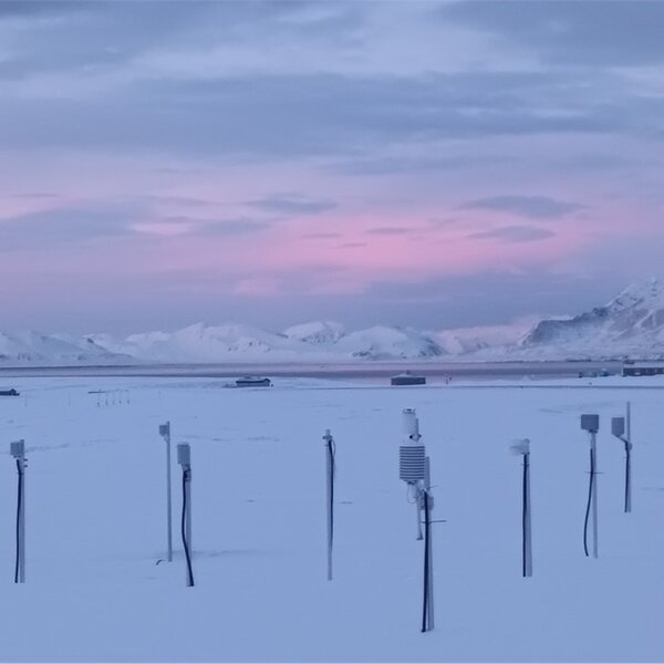 An image showing dawn over the Arctic research station in Norway. In the foreground measuring instruments stand out from the snow and in the background are a range of snow-covered mountains