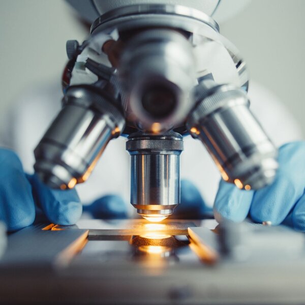 In the centre of the picture is a microscope with 4 lenses. Behind and out of site, sits a scientist with only gloved hands showing either side of the microscope