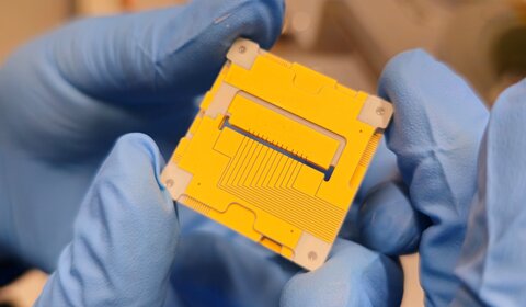 Image showing a pair of blue gloved hands holding the new 3D ion trap, which resembles a yellow square 3 cm by 3 cm