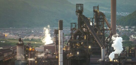 Against a hazy grey sky a large steel plant sits in a valley with white smoke plumes emitted from its large chimney stacks