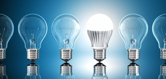 Against a pale blue background is a row of unlit incandescent screw light bulbs with one lit LED bulb