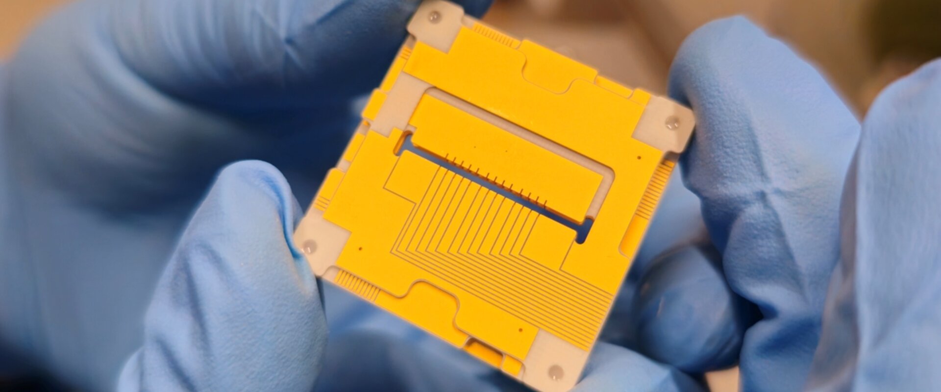 Image showing a pair of blue gloved hands holding the new 3D ion trap, which resembles a yellow square 3 cm by 3 cm