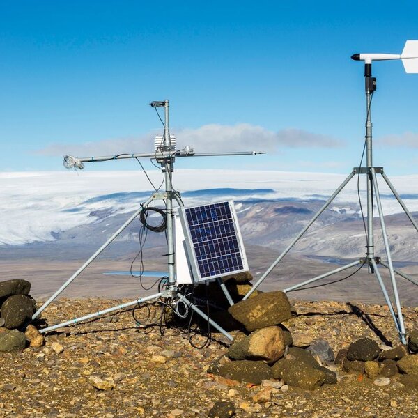 On a rocky ground in the mountains and framed by a blue sky sits a meteorological station. It is composed of a small monitor with a solar panel on the left and on the right a weathervane