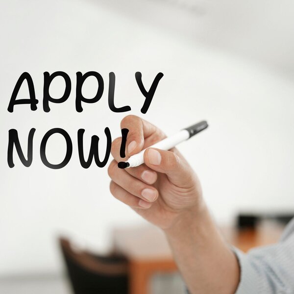 A hand writing “APPLY NOW!” on a whiteboard