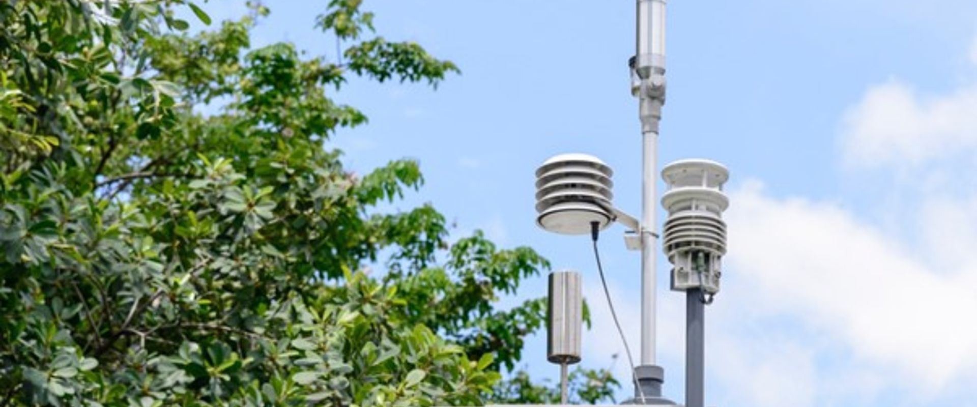An air monitoring station In the centre of the frame, an air monitoring station is attached to a pole. It consists of a grey metal box with a handle pointing towards the viewer and three covered pylons, carrying sampling equipment, protruding from the top, as well as a small silver cylinder. In the background there is the canopy of a full, green tree, and a blue sky filled with soft clouds