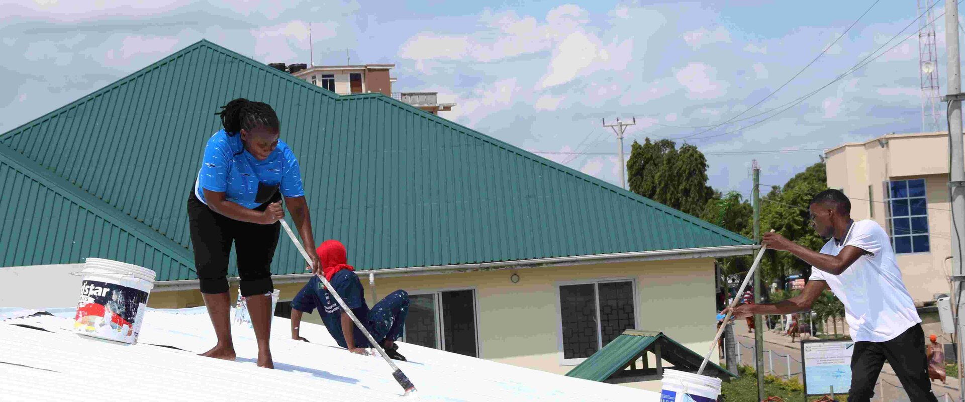 A scene showing roof tops in Tanzania in Africa. On top of the roof are three locals with long handled paint brushes who are painting one of the roofs with bright white paint