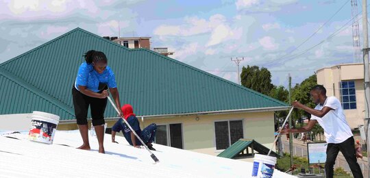 A scene showing roof tops in Tanzania in Africa. On top of the roof are three locals with long handled paint brushes who are painting one of the roofs with bright white paint