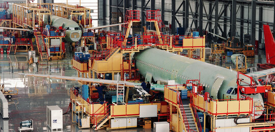 In a vast indoor hanger two commercial aircraft, surrounded by gantries and equipment, are being assembled