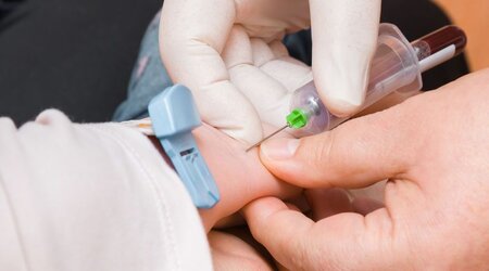 A close-up image of the hands of a doctor with white rubber gloves taking blood out of a young child’s arm with a syringe