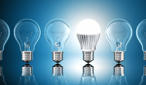 Against a pale blue background is a row of unlit incandescent screw light bulbs with one lit LED bulb