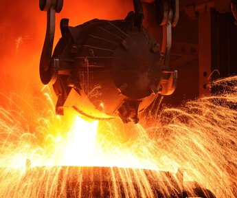 In a factory environment an overhead metal bucket pours orange glowing, molten steel into a vessel below