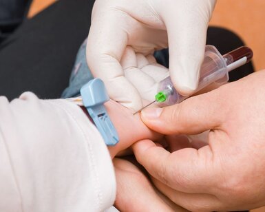A close-up image of the hands of a doctor with white rubber gloves taking blood out of a young child’s arm with a syringe