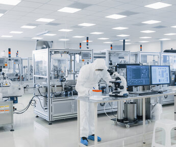 In a high-tech laboratory filled with sterile cabinets a worker in protective overalls and mask performs research using a microscope
