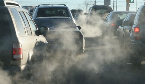 A photograph of a number of cars stopped at a traffic slight. The image is hazy with exhaust fumes coming from the vehicles’ exhausts