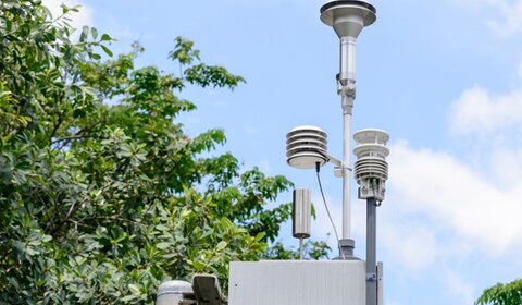 In the centre of the frame, an air monitoring station is attached to a pole. It consists of a grey metal box with a handle pointing towards the viewer and three covered pylons, carrying sampling equipment, protruding from the top, as well as a small silver cylinder. In the background there is the canopy of a full, green tree, and a blue sky filled with soft clouds