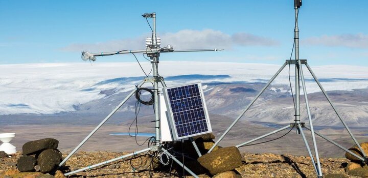 On a rocky ground in the mountains and framed by a blue sky sits a meteorological station. It is composed of a small monitor with a solar panel on the left and on the right a weathervane